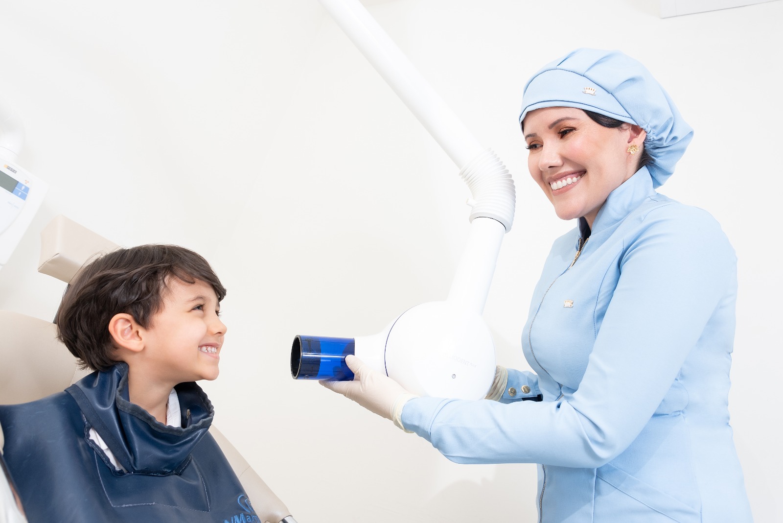 Profissional da Sigma posiciona o aparelho de raio-x ao lado de um menino sentado na cadeira, os dois sorrindo.